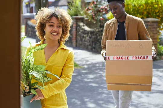woman holding a potted plant, man holding a box that says Fragile, entering into a house